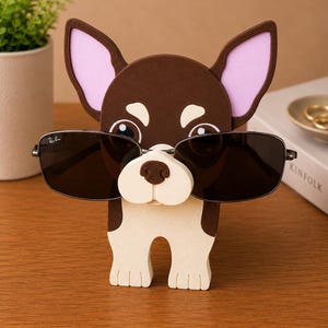 Puede incluir: Un soporte para gafas con forma de chihuahua marr&oacute;n y blanco con orejas forradas de rosa. El soporte sostiene un par de gafas de sol negras con montura plateada. Las gafas de sol tienen el logotipo de Ray-Ban.