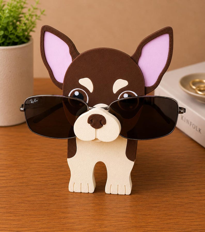 Puede incluir: Un soporte para gafas con forma de chihuahua marr&oacute;n y blanco con orejas forradas de rosa. El soporte sostiene un par de gafas de sol negras con montura plateada. Las gafas de sol tienen el logotipo de Ray-Ban.
