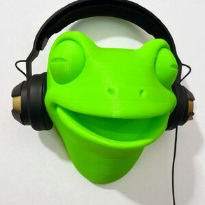 May include: A vibrant green frog-shaped headphone holder with a smiling face. It features black headphones with gold detailing. A black headphone cable hangs down. The holder is mounted on a white wall.