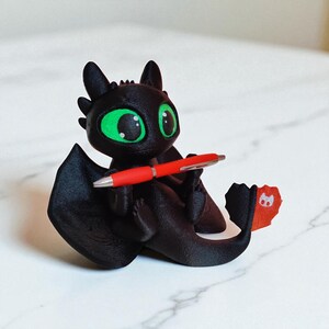 May include: A black dragon figurine with large green eyes, holding a red pen. The dragon has large wings and a tail with a red and orange fin. The figurine is sitting on a white marble surface.