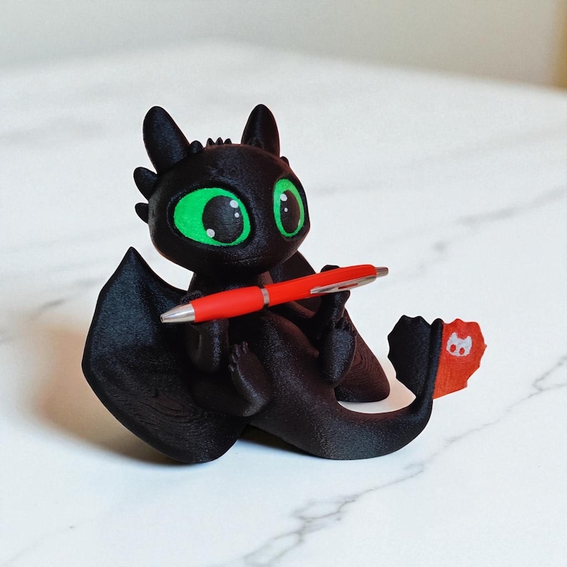 May include: A black dragon figurine with large green eyes, holding a red pen. The dragon has large wings and a tail with a red and orange fin. The figurine is sitting on a white marble surface.