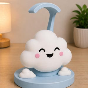 May include: A whimsical cloud-shaped night light in white and light blue. The cloud has a smiling face with pink cheeks and sits on a light blue base. A curved handle extends from the top. Decorative and functional.