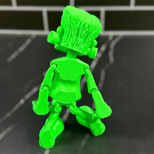 May include: A green articulated toy figure of Frankenstein's monster. The figure is posed with its arms bent and its legs crossed. The figure is made of a textured plastic.