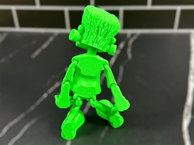 May include: A green articulated toy figure of Frankenstein's monster. The figure is posed with its arms bent and its legs crossed. The figure is made of a textured plastic.