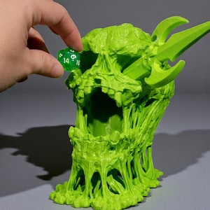 May include: A lime green skull-shaped dice tower with a sword-like element. A hand is placing a green die into the tower. The tower has a textured, dripping design. The dice shows the number 16.