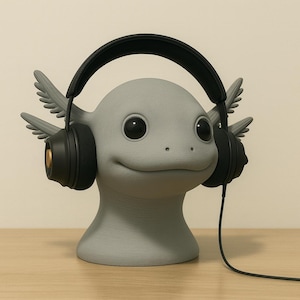 Può includere: Un supporto per cuffie a forma di axolotl grigio con grandi occhi neri e branchie piumate. Il supporto ha cuffie nere con dettagli dorati. L'axolotl ha un'espressione sorridente ed è su una superficie di legno.