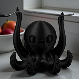 May include: A black octopus-shaped spoon rest. The octopus has a round head, large eyes, and eight tentacles. The spoon rest is holding a silver spoon. The octopus is sitting on a white surface.