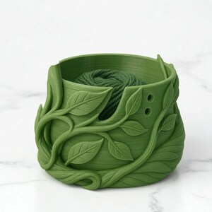 May include: A green yarn bowl with a leaf design. The bowl is filled with a ball of green yarn. The exterior of the bowl is decorated with detailed, three-dimensional leaves and vines. The bowl has two small holes on the side.