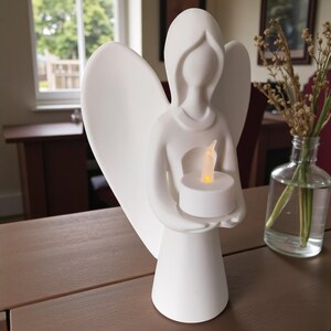 May include: A white angel figurine with outstretched wings, holding a lit LED candle. The angel has a simple, elegant design and is placed on a brown wooden surface. The background includes a window, a table, and a vase with dried flowers.