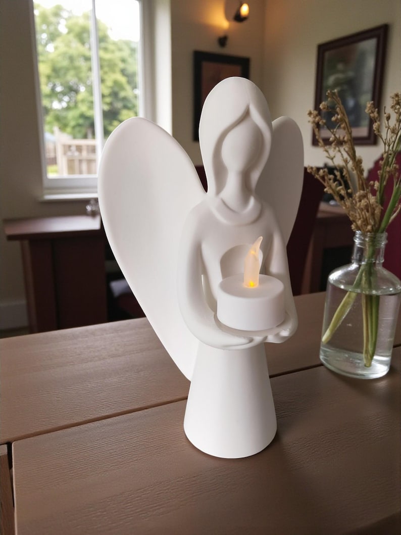 May include: A white angel figurine with outstretched wings, holding a lit LED candle. The angel has a simple, elegant design and is placed on a brown wooden surface. The background includes a window, a table, and a vase with dried flowers.