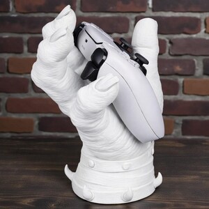 May include: A white, hand-shaped gaming controller stand. The stand is holding a white and black gaming controller. The base of the stand has a decorative design. The background is a brick wall and a wooden surface.