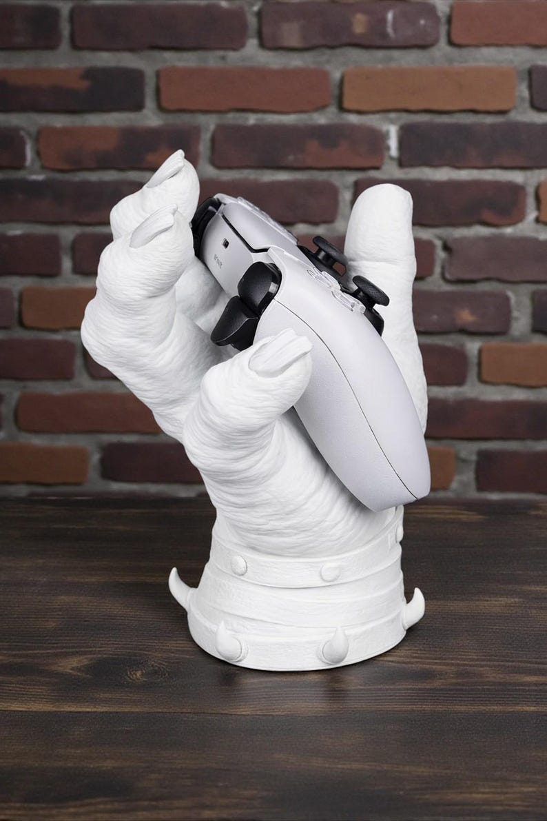 May include: A white, hand-shaped gaming controller stand. The stand is holding a white and black gaming controller. The base of the stand has a decorative design. The background is a brick wall and a wooden surface.