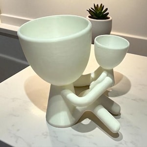 Op de afbeelding: Een witte keramische plantenbak in de vorm van een persoon die een kleinere plantenbak vasthoudt. De grotere plantenbak heeft een brede, open komvorm, terwijl de kleinere in de armen van de figuur wordt gehouden. Een kleine vetplant in een witte pot staat op de achtergrond.