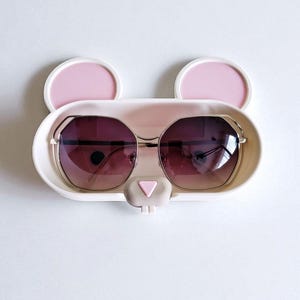 May include: A white and pink sunglasses holder in the shape of a bear's face, with two pink ears and a pink nose. The holder contains a pair of sunglasses with a gold frame and gradient lenses.