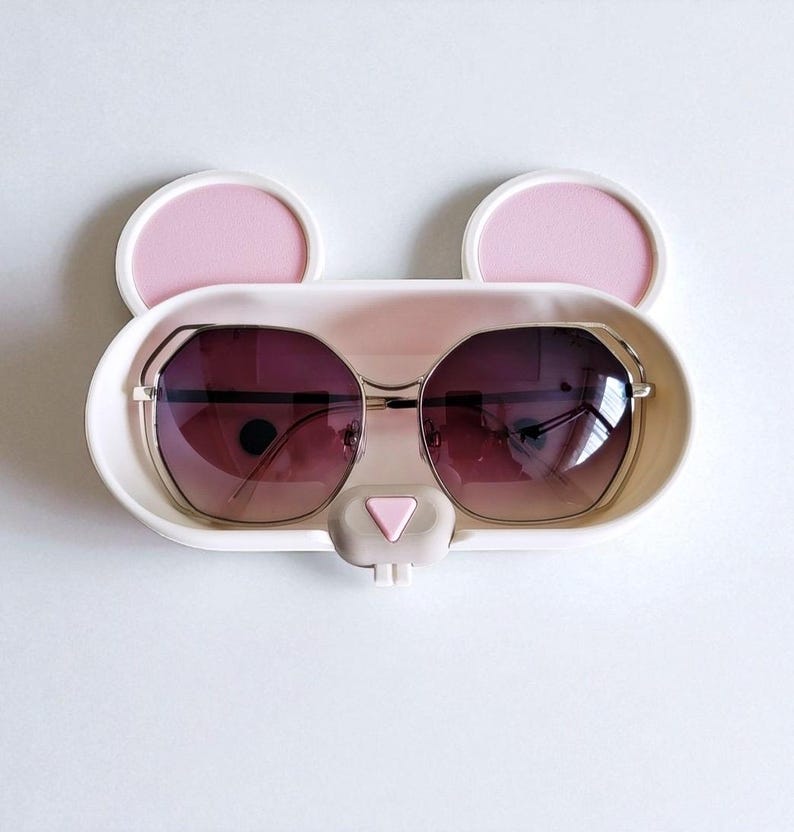 May include: A white and pink sunglasses holder in the shape of a bear's face, with two pink ears and a pink nose. The holder contains a pair of sunglasses with a gold frame and gradient lenses.
