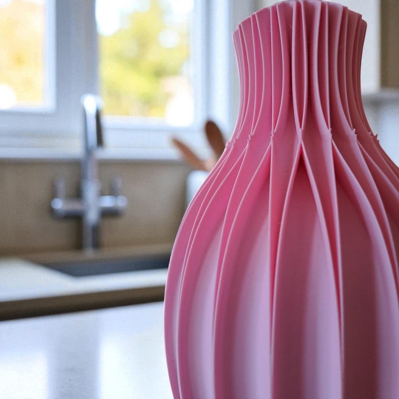 May include: A close-up of a pink vase with a unique, pleated design. The vase has a wide base that tapers to a narrow neck. The pink color is consistent throughout the vase, and the pleats create a textured surface. The vase is likely made of ceramic or plastic.
