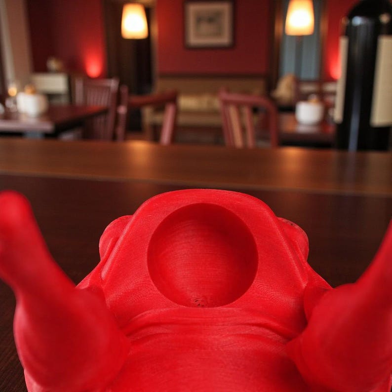 May include: A vibrant red wine bottle holder, 3D-printed with a central circular opening and two extended arms. The background shows a restaurant setting with tables, chairs, and warm lighting.