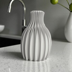 May include: A white, decorative vase with a unique, pleated design. The vase has a bulbous body and a narrow neck, with vertical pleats creating a textured surface. The vase is made of a solid, opaque material and is suitable for home decor.