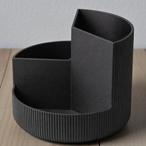K&ouml;nnte beinhalten: Ein schwarzer Schreibtisch-Organizer mit halbkreisf&ouml;rmiger Basis und drei F&auml;chern. Der Organizer hat ein strukturiertes, geriffeltes Design an der Basis und einer Seite. Die F&auml;cher sind unterschiedlich gro&szlig; und geformt.