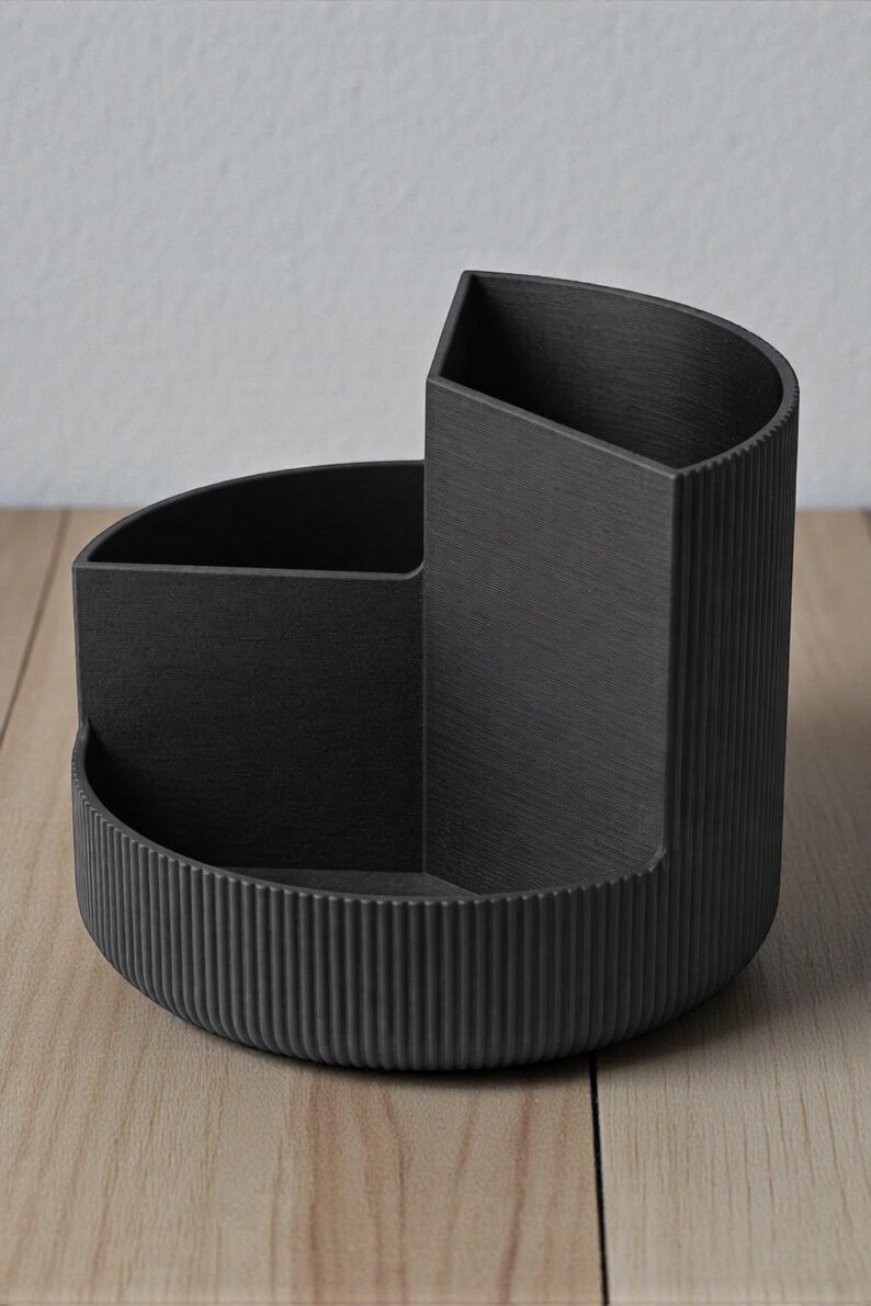 K&ouml;nnte beinhalten: Ein schwarzer Schreibtisch-Organizer mit halbkreisf&ouml;rmiger Basis und drei F&auml;chern. Der Organizer hat ein strukturiertes, geriffeltes Design an der Basis und einer Seite. Die F&auml;cher sind unterschiedlich gro&szlig; und geformt.