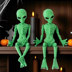 May include: Two articulated green alien figures with large black eyes sit on a shelf. The figures have detailed rib cages and are posed with their legs dangling. The shelf is decorated with pumpkins, candles, and a spider web, creating a Halloween theme.