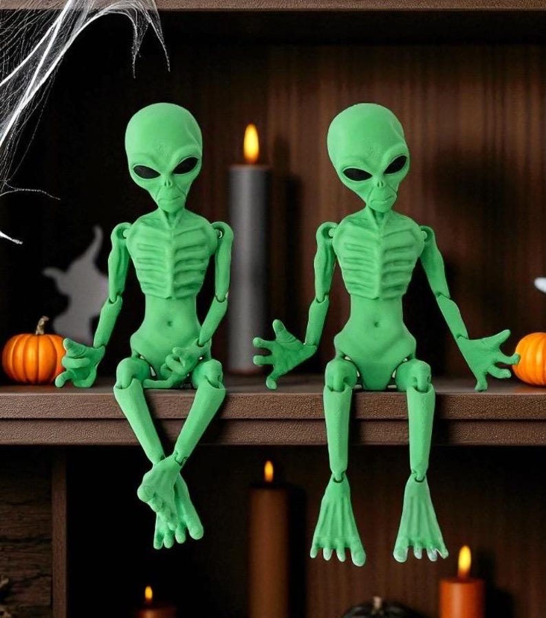 May include: Two articulated green alien figures with large black eyes sit on a shelf. The figures have detailed rib cages and are posed with their legs dangling. The shelf is decorated with pumpkins, candles, and a spider web, creating a Halloween theme.
