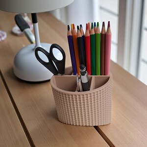 May include: A beige desk organizer with a textured pattern, holding colored pencils, a pen, and scissors. The pencils are various colors, including purple, red, and green. A white lamp is in the background.