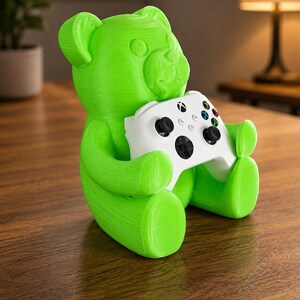 May include: A bright green bear-shaped gaming controller holder. The bear is holding a white Xbox controller with black buttons and colorful directional buttons. The holder is made of a textured material and sits on a wooden surface.