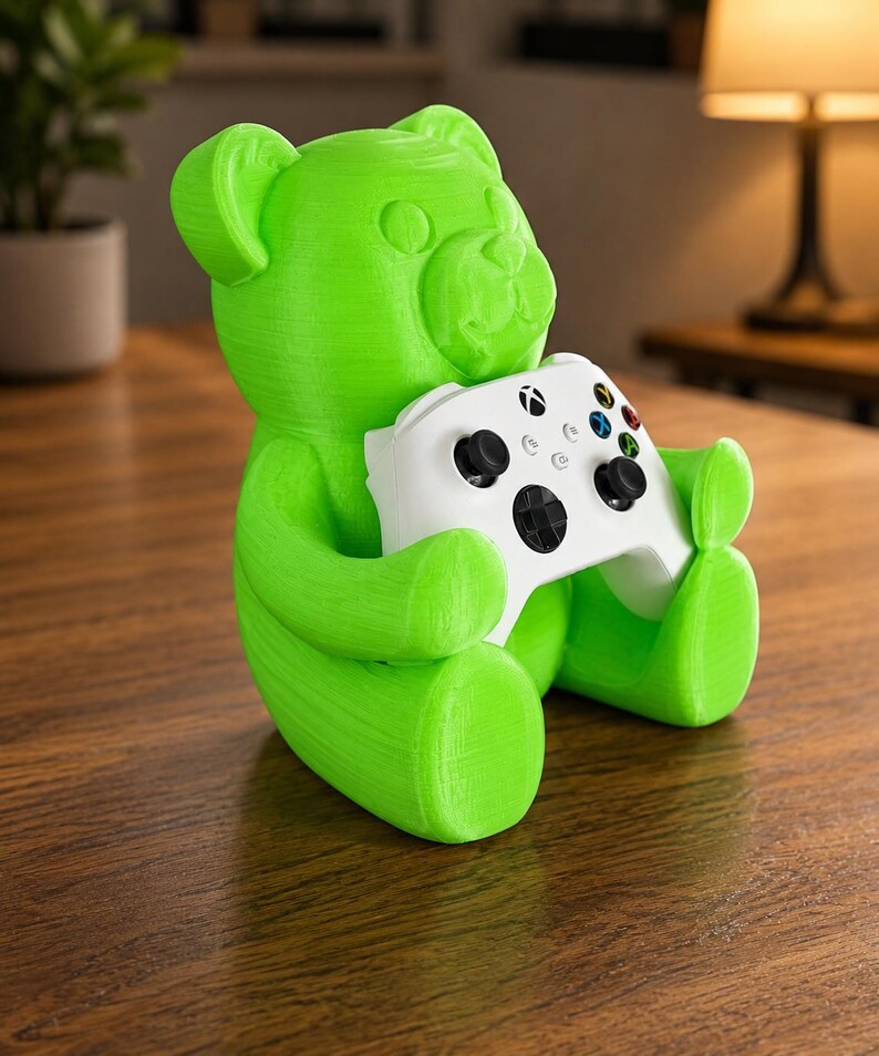 May include: A bright green bear-shaped gaming controller holder. The bear is holding a white Xbox controller with black buttons and colorful directional buttons. The holder is made of a textured material and sits on a wooden surface.