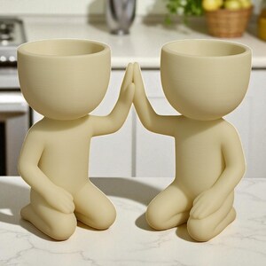 May include: Two beige human-shaped planters, kneeling with hands touching in a high-five gesture. Each planter has a rounded head with a bowl-shaped opening for plants. The planters are on a white marble surface.