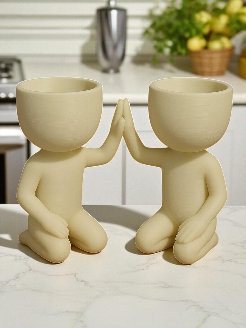 May include: Two beige human-shaped planters, kneeling with hands touching in a high-five gesture. Each planter has a rounded head with a bowl-shaped opening for plants. The planters are on a white marble surface.