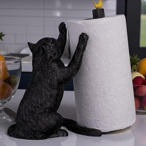 May include: A black cat-shaped paper towel holder. The cat is sculpted in a playful pose, reaching up towards the white paper towel roll. A small yellow bird sits atop the holder. The paper towel roll has a floral pattern.