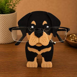 May include: A black and tan Rottweiler dog-shaped eyeglass holder. The holder is made of wood and has a black frame holding a pair of eyeglasses. The dog's face is tan with black accents, and the eyeglasses are clear.