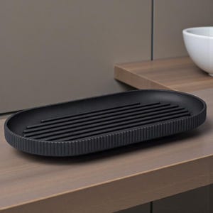 May include: A matte black, oval-shaped soap dish with a ribbed exterior and a series of parallel bars across the center. The dish sits on a wooden surface, with a neutral-toned background. The design is minimalist and modern.