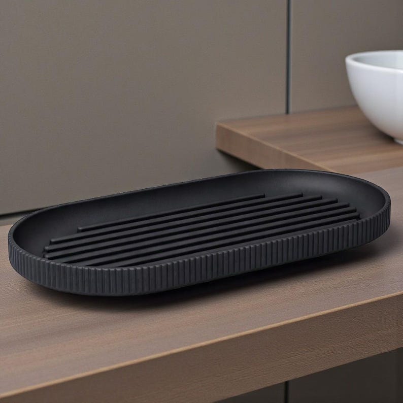 May include: A matte black, oval-shaped soap dish with a ribbed exterior and a series of parallel bars across the center. The dish sits on a wooden surface, with a neutral-toned background. The design is minimalist and modern.
