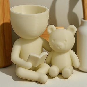 May include: A cream-coloured planter in the shape of a person reading a book, with a small teddy bear beside it. The planter has a large head with a bowl-shaped opening. The bear is also cream-coloured.