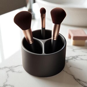 Puede incluir: Un soporte para brochas de maquillaje gris oscuro con tres compartimentos, que contiene tres brochas de maquillaje. Las brochas tienen cerdas marrones y mangos de oro rosa. Una peque&ntilde;a esponja rosa est&aacute; en el fondo.