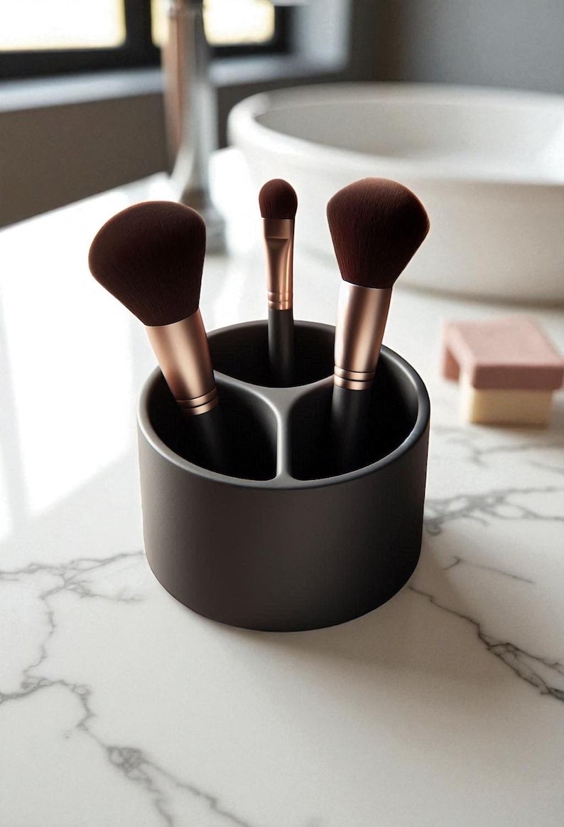 Puede incluir: Un soporte para brochas de maquillaje gris oscuro con tres compartimentos, que contiene tres brochas de maquillaje. Las brochas tienen cerdas marrones y mangos de oro rosa. Una peque&ntilde;a esponja rosa est&aacute; en el fondo.