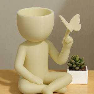 Puede incluir: Una figura de color amarillo claro de una persona sentada, con una mariposa en un dedo. La cabeza es una maceta. Una peque&ntilde;a planta suculenta en una maceta cuadrada blanca est&aacute; cerca. La figura est&aacute; sobre una superficie de madera.