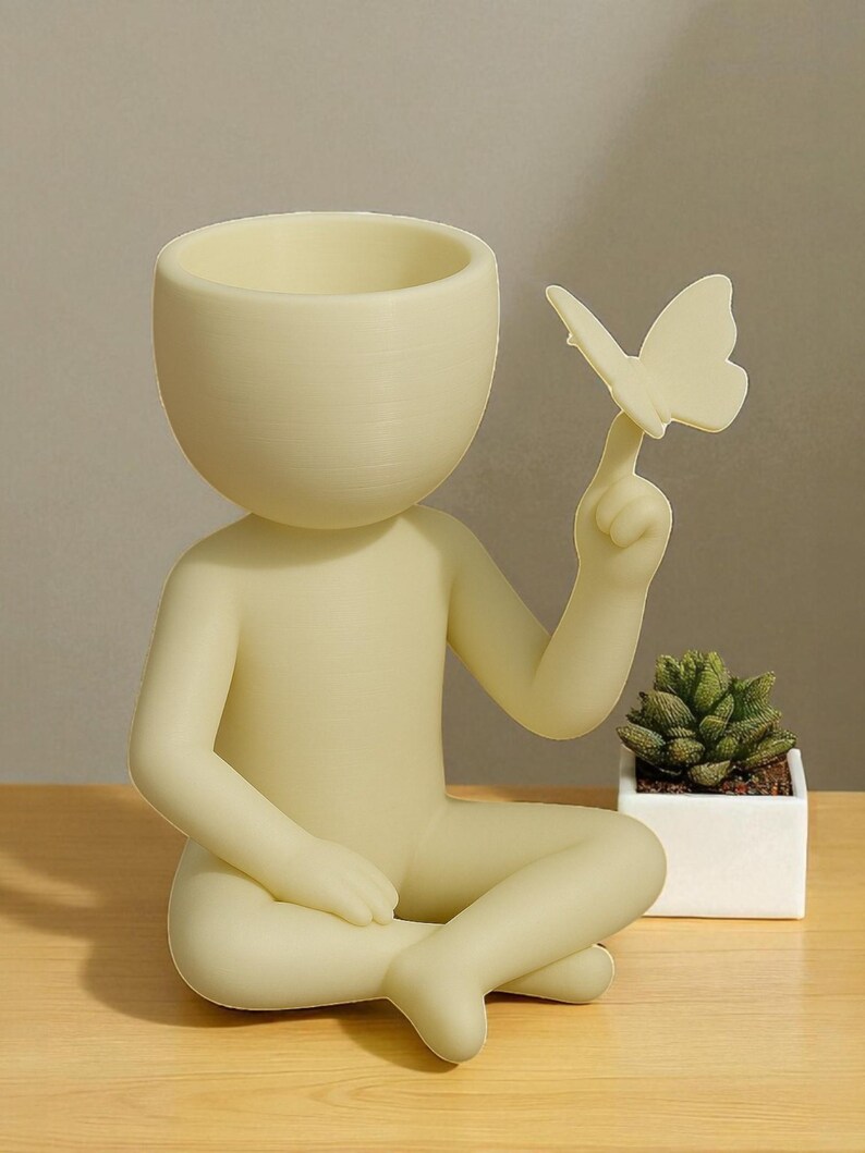 Puede incluir: Una figura de color amarillo claro de una persona sentada, con una mariposa en un dedo. La cabeza es una maceta. Una peque&ntilde;a planta suculenta en una maceta cuadrada blanca est&aacute; cerca. La figura est&aacute; sobre una superficie de madera.