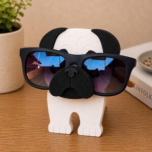 May include: A white and black pug-shaped glasses holder with a pair of black sunglasses. The sunglasses have blue and purple lenses. The holder is made of wood and sits on a wooden surface.