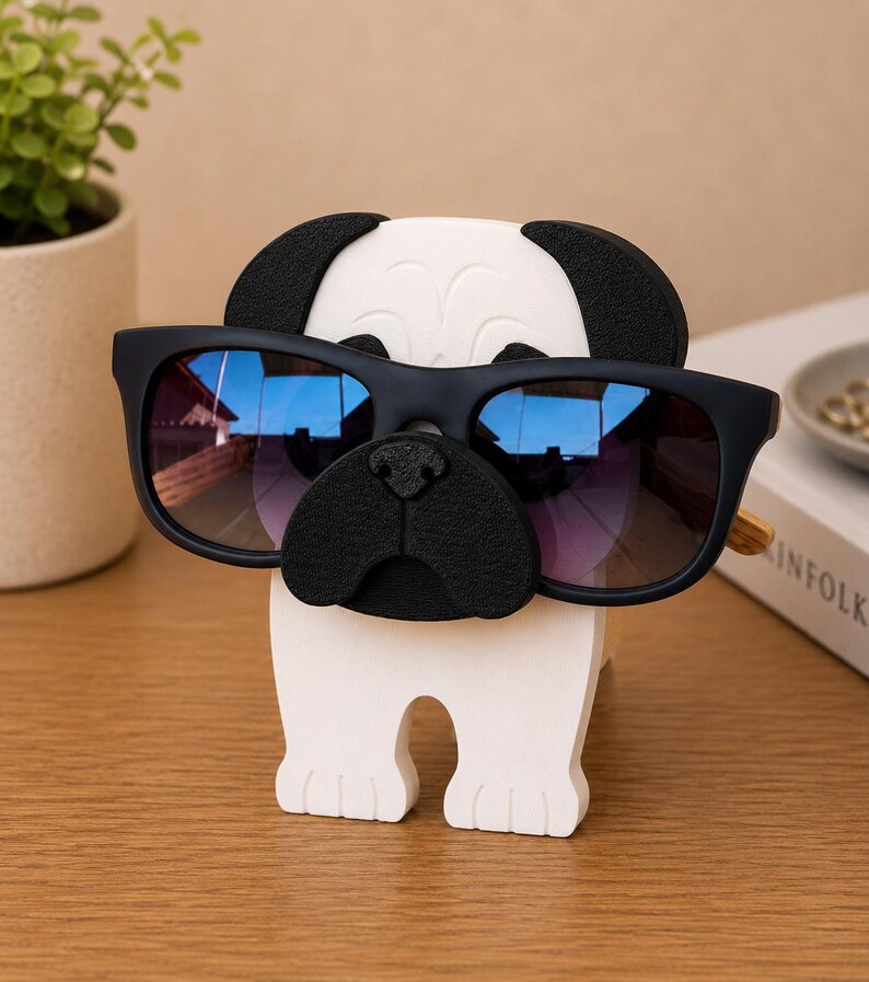 May include: A white and black pug-shaped glasses holder with a pair of black sunglasses. The sunglasses have blue and purple lenses. The holder is made of wood and sits on a wooden surface.
