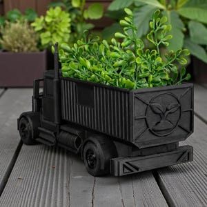 K&ouml;nnte beinhalten: Ein schwarzer, LKW-f&ouml;rmiger Pflanzk&uuml;bel, gef&uuml;llt mit gr&uuml;nen Pflanzen. Der Pflanzk&uuml;bel hat ein detailliertes Design, einschlie&szlig;lich R&auml;dern, Kabine und Containerbett. Das Containerbett hat ein kreisf&ouml;rmiges Emblem. Der Pflanzk&uuml;bel steht auf einer Holzoberfl&auml;che.