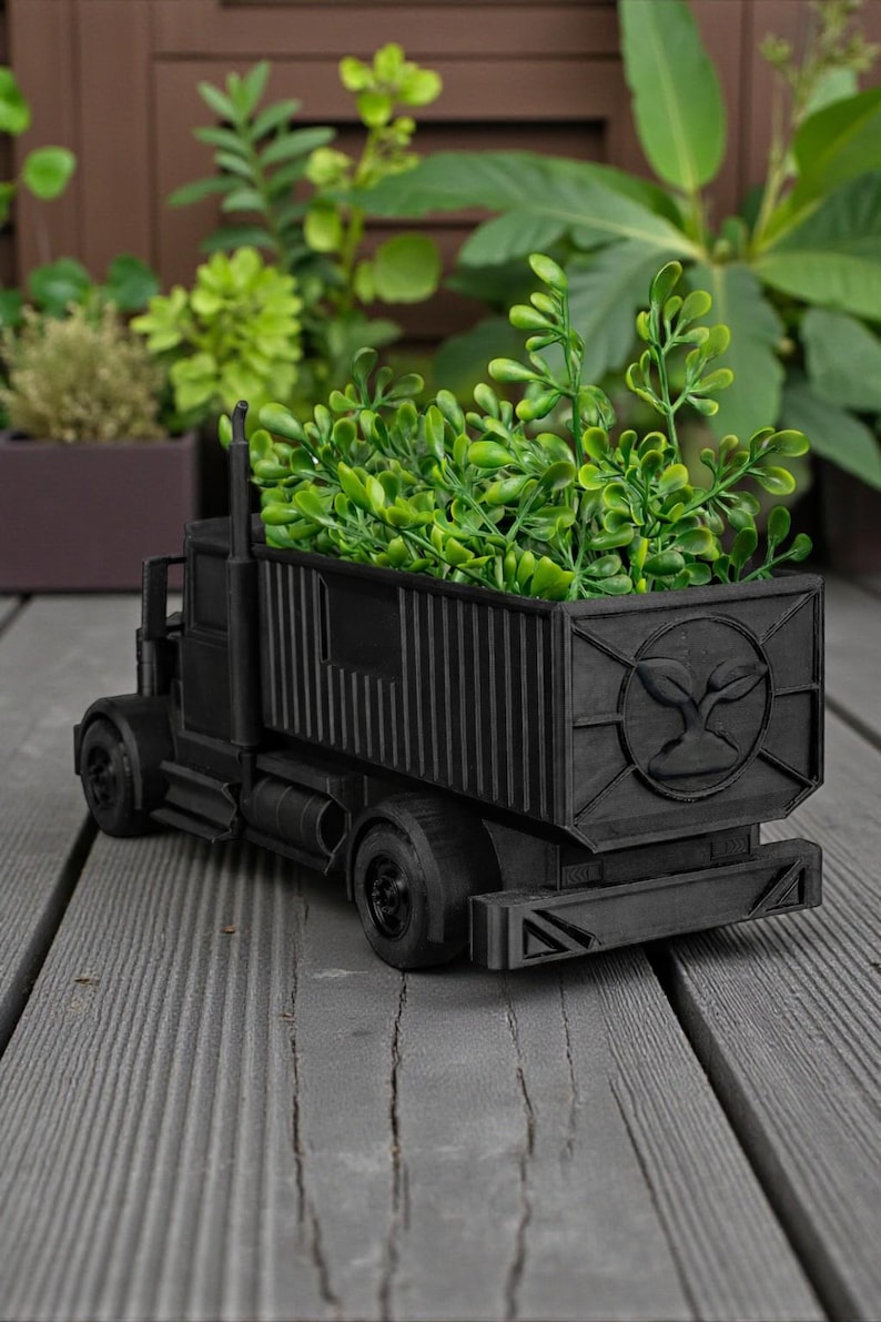 K&ouml;nnte beinhalten: Ein schwarzer, LKW-f&ouml;rmiger Pflanzk&uuml;bel, gef&uuml;llt mit gr&uuml;nen Pflanzen. Der Pflanzk&uuml;bel hat ein detailliertes Design, einschlie&szlig;lich R&auml;dern, Kabine und Containerbett. Das Containerbett hat ein kreisf&ouml;rmiges Emblem. Der Pflanzk&uuml;bel steht auf einer Holzoberfl&auml;che.
