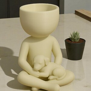 May include: A cream-colored figurine of a person seated with legs crossed, cradling a sleeping puppy. The figure has a large, open bowl-shaped head, suitable for a small plant. A small potted succulent sits nearby, adding a touch of greenery.