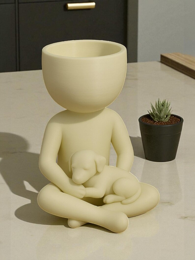 May include: A cream-colored figurine of a person seated with legs crossed, cradling a sleeping puppy. The figure has a large, open bowl-shaped head, suitable for a small plant. A small potted succulent sits nearby, adding a touch of greenery.