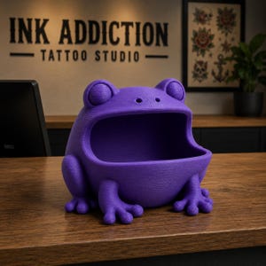May include: A purple frog-shaped container with an open mouth, sitting on a wooden surface. The container has detailed features, including eyes and feet. The background includes the text "INK ADDICTION TATTOO STUDIO".