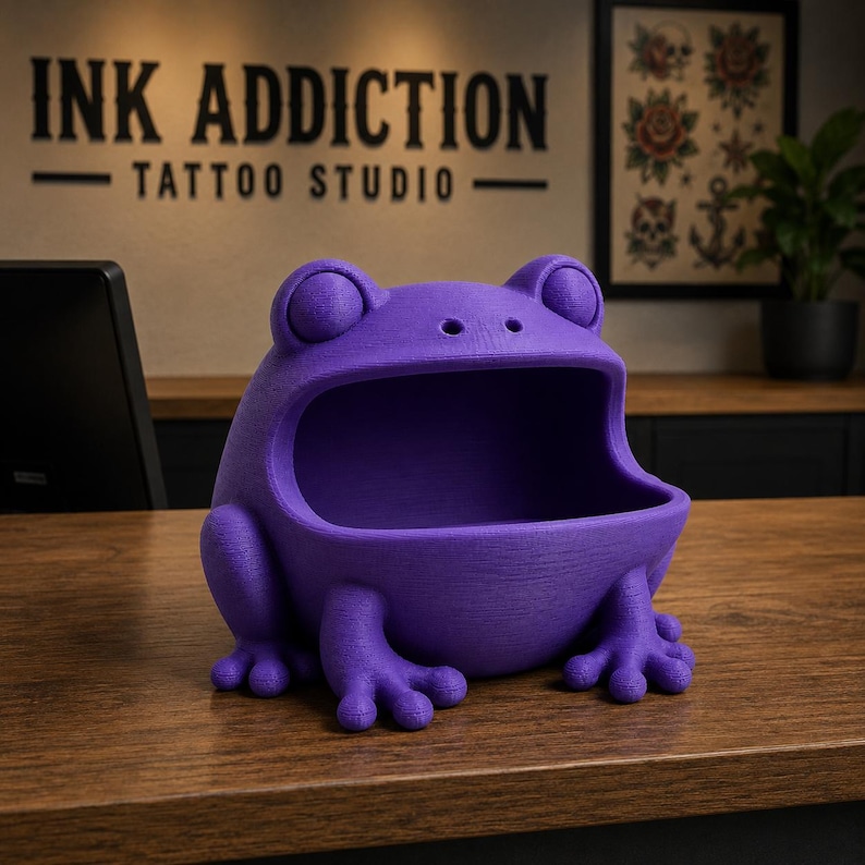 May include: A purple frog-shaped container with an open mouth, sitting on a wooden surface. The container has detailed features, including eyes and feet. The background includes the text "INK ADDICTION TATTOO STUDIO".