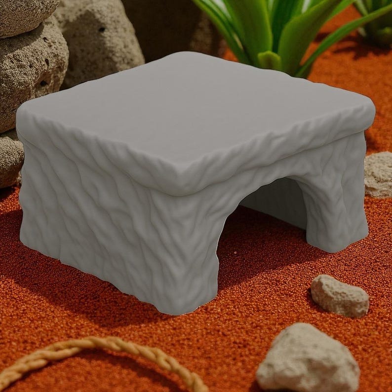 May include: A gray, cave-like reptile hideaway with a textured exterior. The hideaway has a rectangular top and an arched opening. The product is designed to provide shelter for small pets. The hideaway is sitting on a bed of orange sand.
