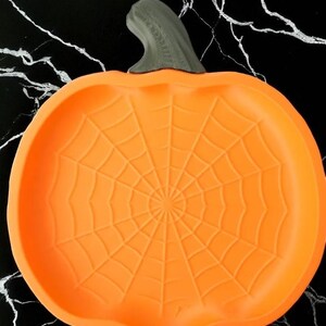 May include: Orange pumpkin-shaped serving dish with a gray stem. The dish features a spiderweb design in the center, perfect for Halloween. The dish is made of a flexible material.